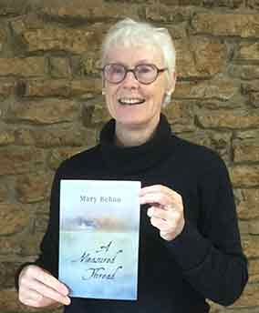 Photo shows author Mary Behan and her book titled A Measured Thread. Mary is white, has short white hair, and wears round, brown glasses.