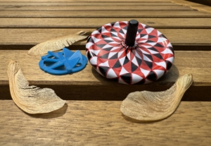 Image shows two spinning tops, a flat blue spinner and a white, black, and white spinner on a table with some maple helicopter seeds.